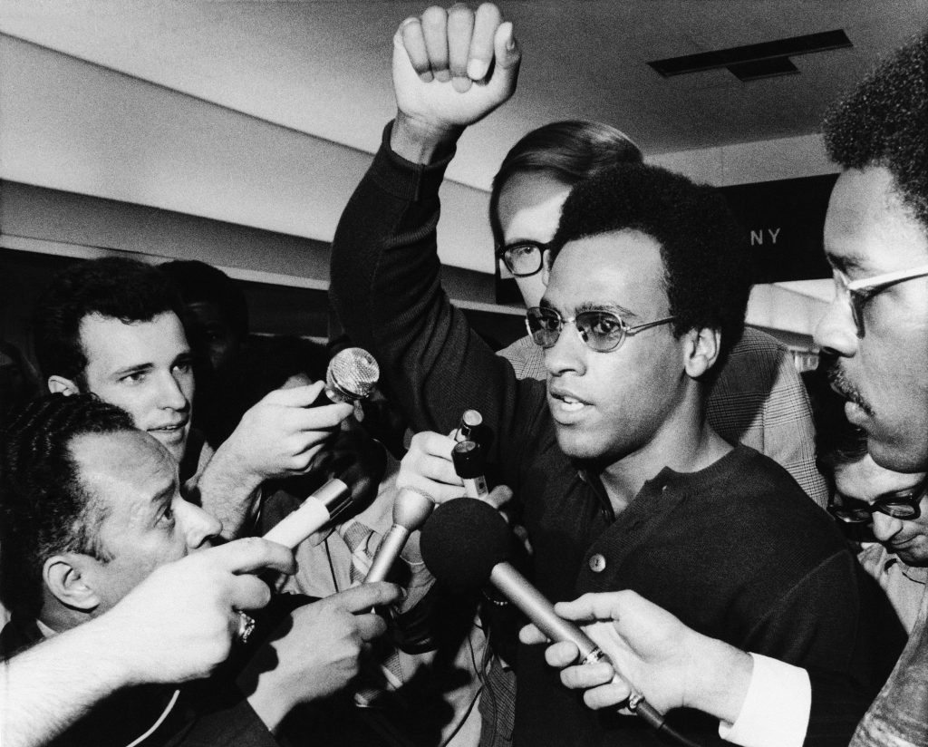 Huey P. Newton, Black Panther party minister of defense, raises his arm as he is literally surrounded by newsmen and others at Philadelphias International Airport on Friday, Sept. 4, 1970 in Philadelphia upon his arrival for a three-day convention. Man at right is not identified. (AP Photo/Rusty Kennedy)